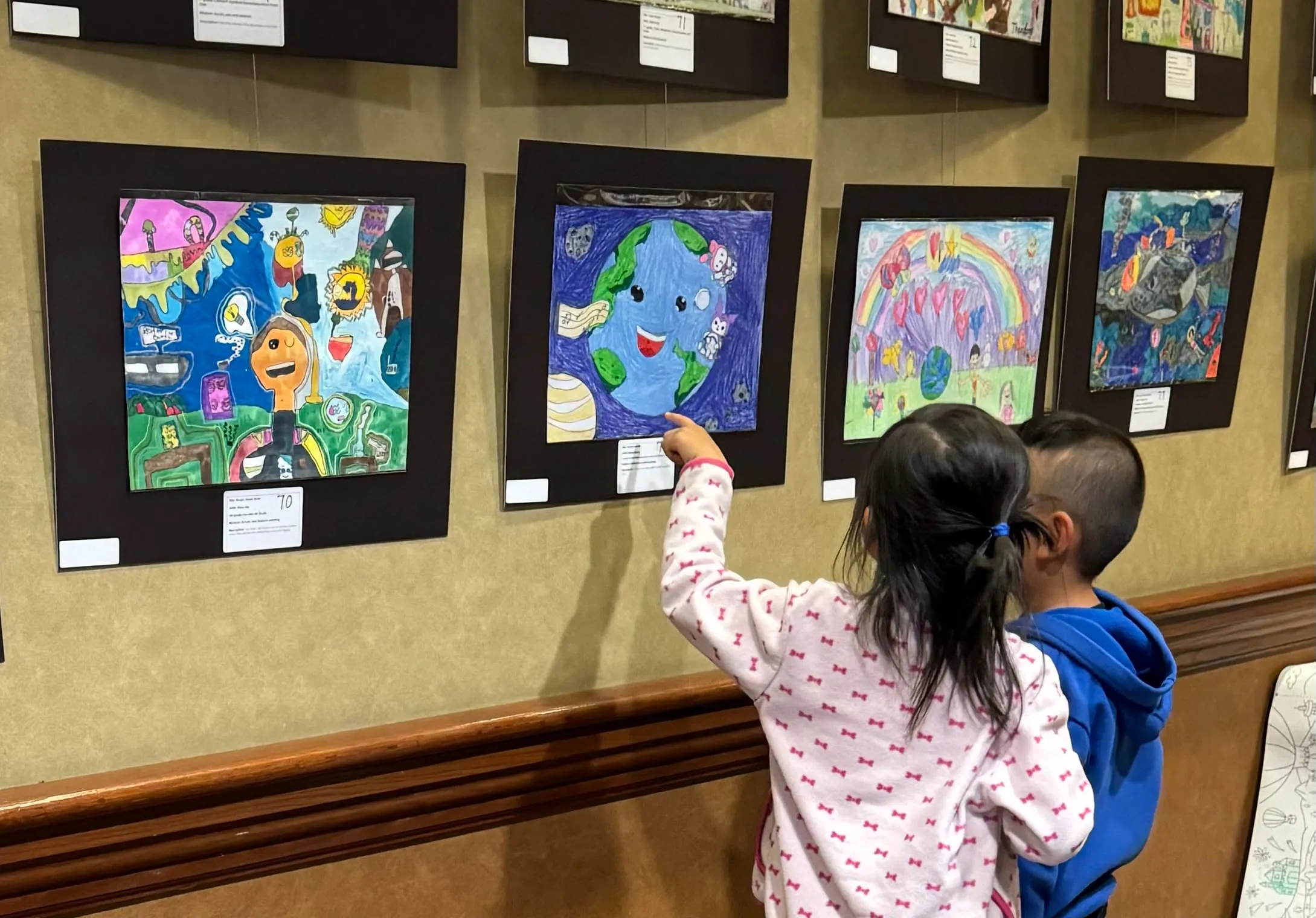 Two young children reach up to point at a painting of the Earth on the Harmony of Hearts exhibition wall.